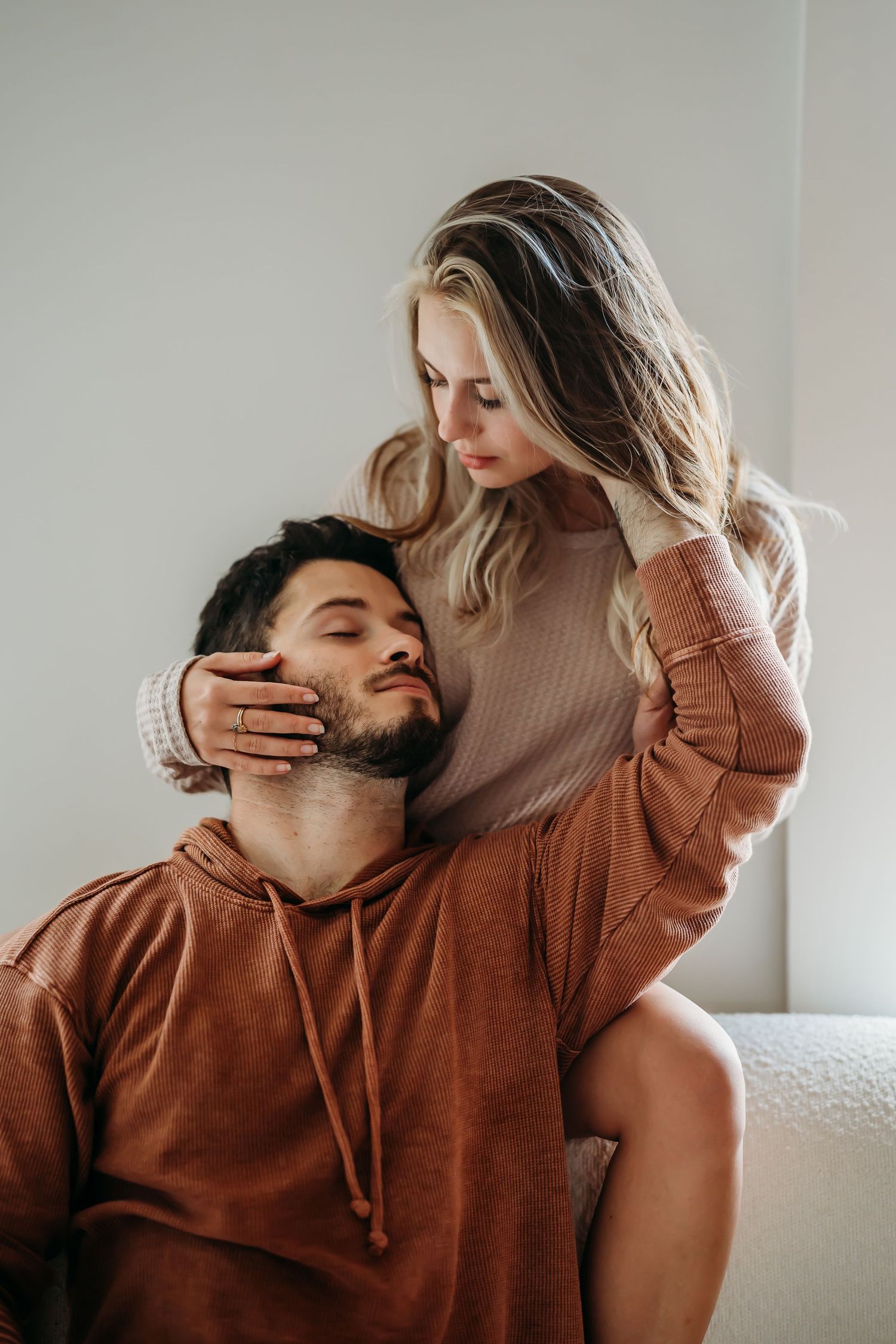 A woman gently cradling a bearded mans face while sitting behind him on a white sofa.
