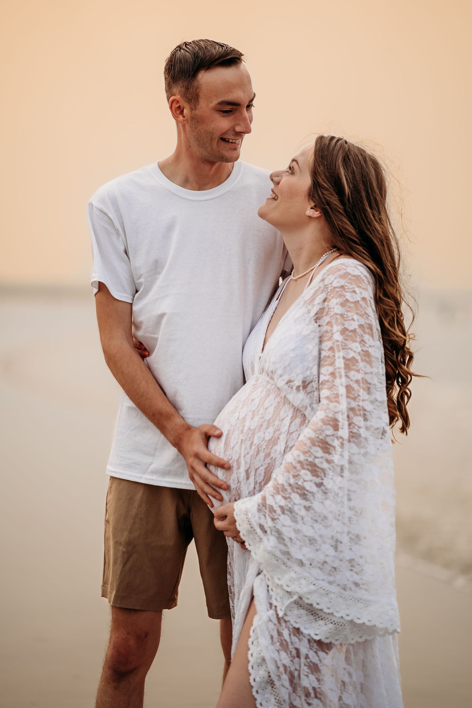 Happy expectant couple holding pregnant belly together on sandy beach at golden hour.