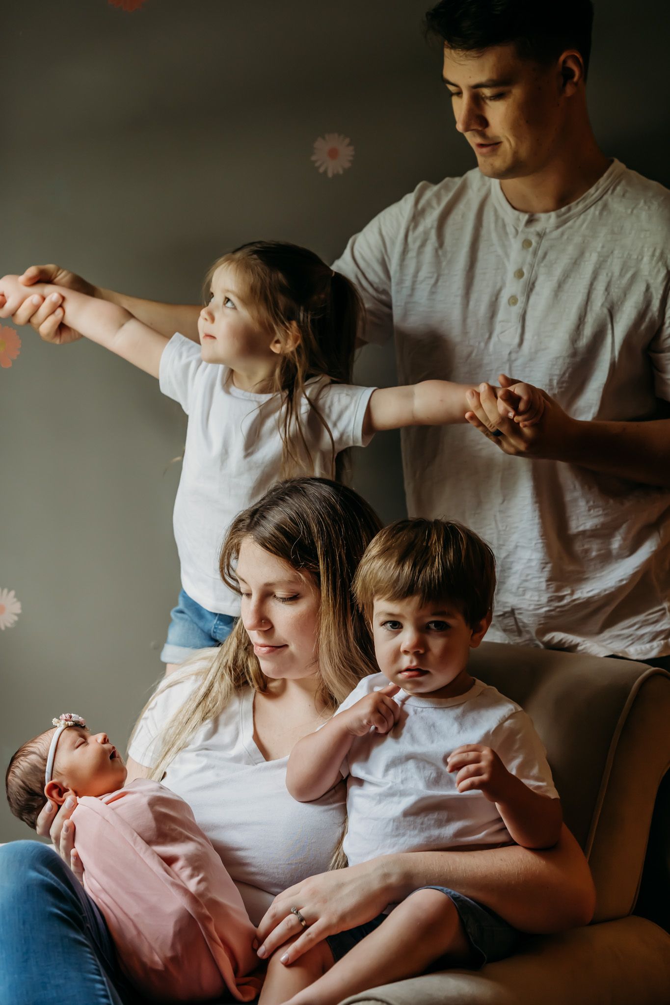 A young family of five poses indoors, with parents holding a newborn, toddler, and playful girl reaching out.