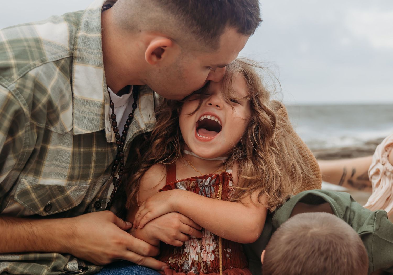 Father kissing laughing little girl on the cheek at the beach, sharing a joyful family moment outdoors.