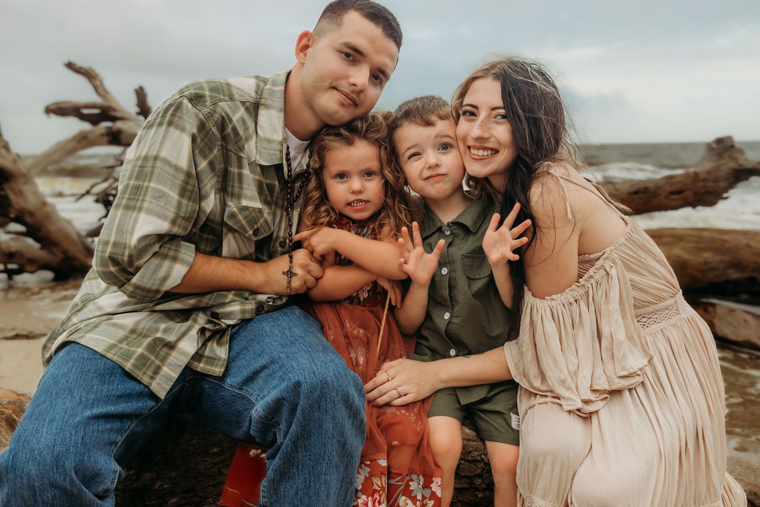 Happy family of four posing together on a driftwood beach, smiling couple with two young children.