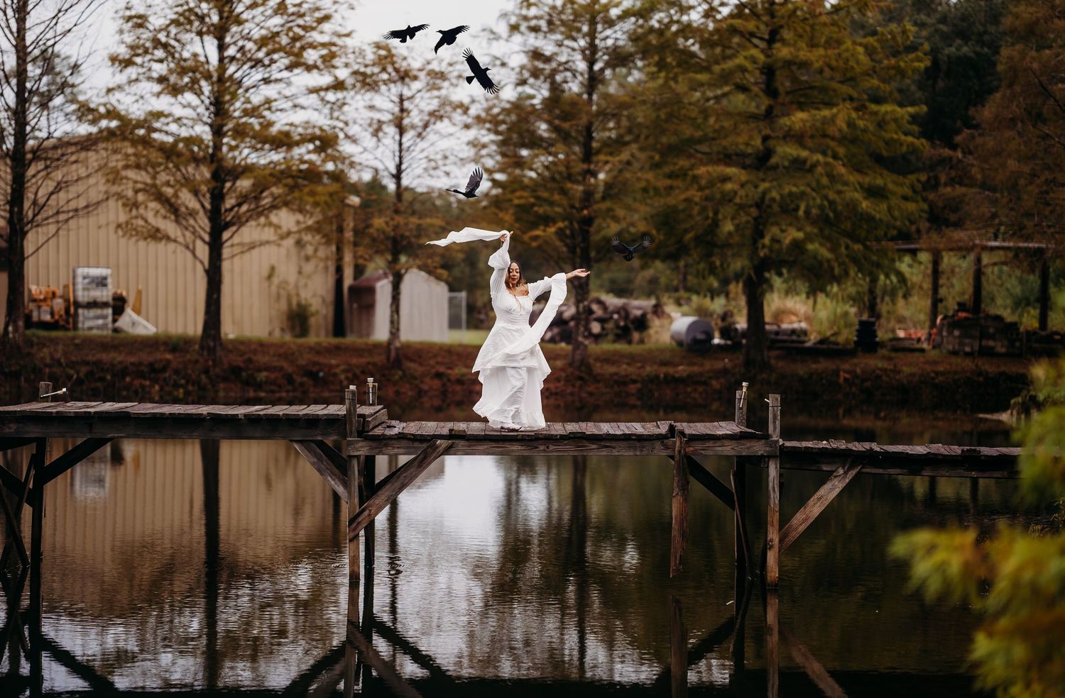 Bride in flowing white dress twirls on a rustic wooden dock with birds flying overhead.