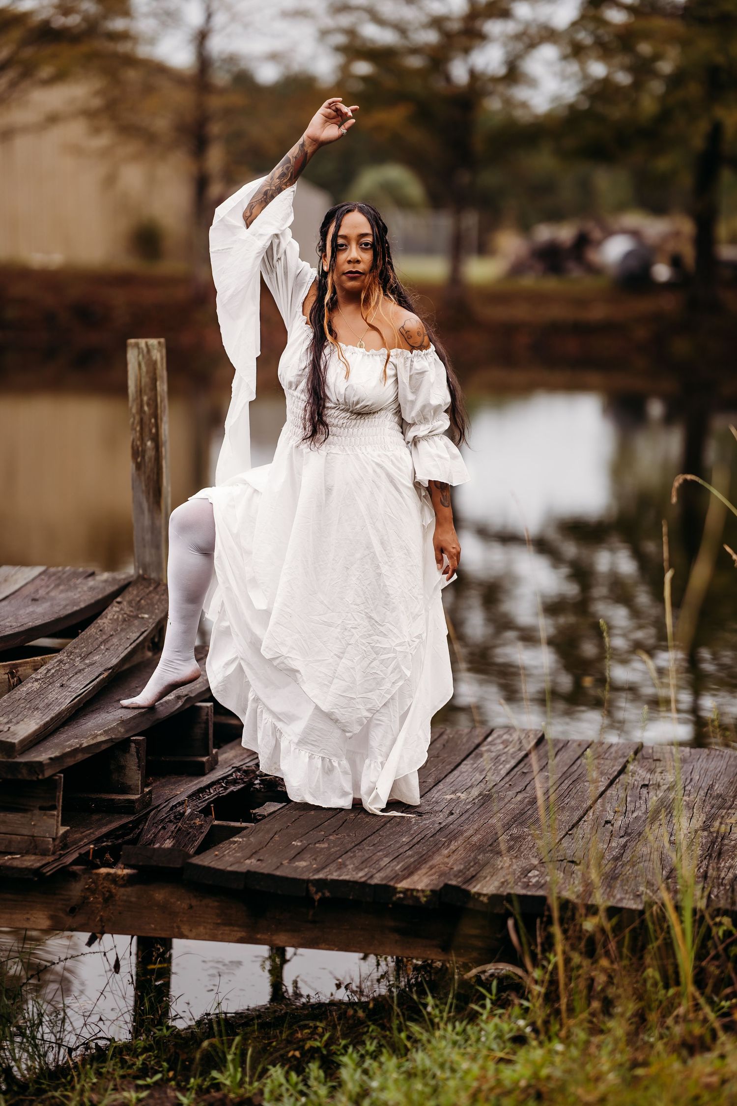 Black woman in flowing white dress raises arm gracefully on weathered wooden pier near pond.