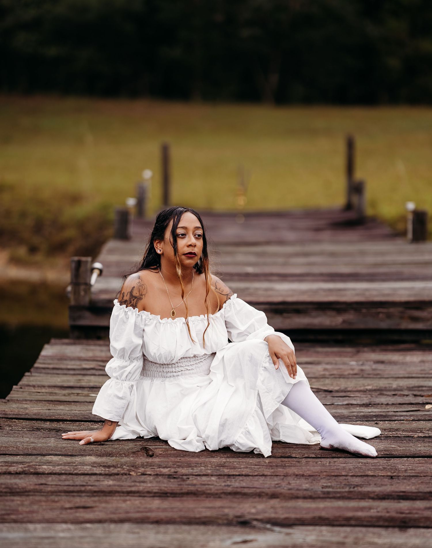 Woman in white off-shoulder dress and white stockings sitting on wooden dock outdoors.