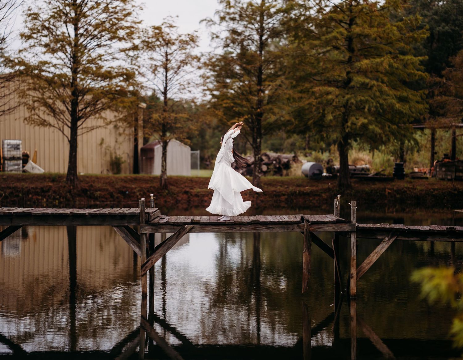 Bride in white gown and veil walking along wooden dock reflected in calm pond with autumn trees.