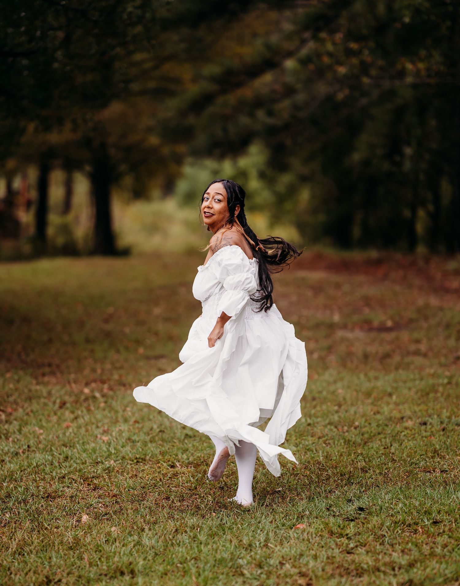 Woman in flowing white off-shoulder dress twirls joyfully in green meadow surrounded by autumn trees.