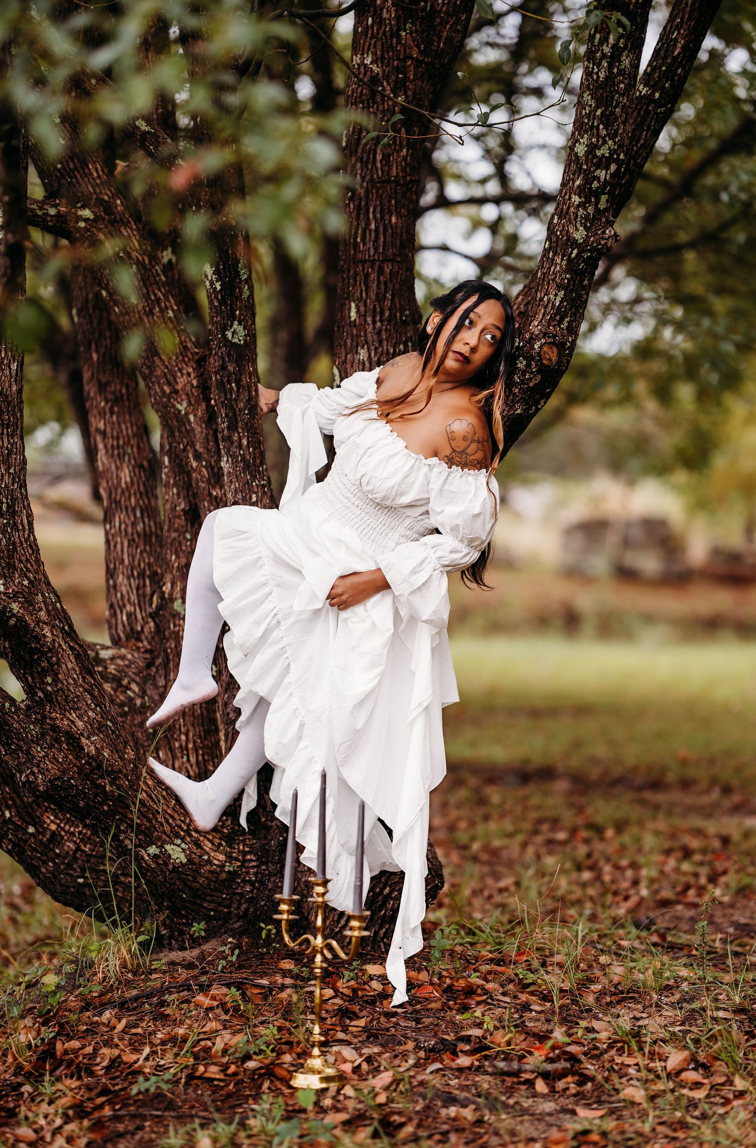 Woman in white ruffled dress posing dramatically against tree in autumn park.