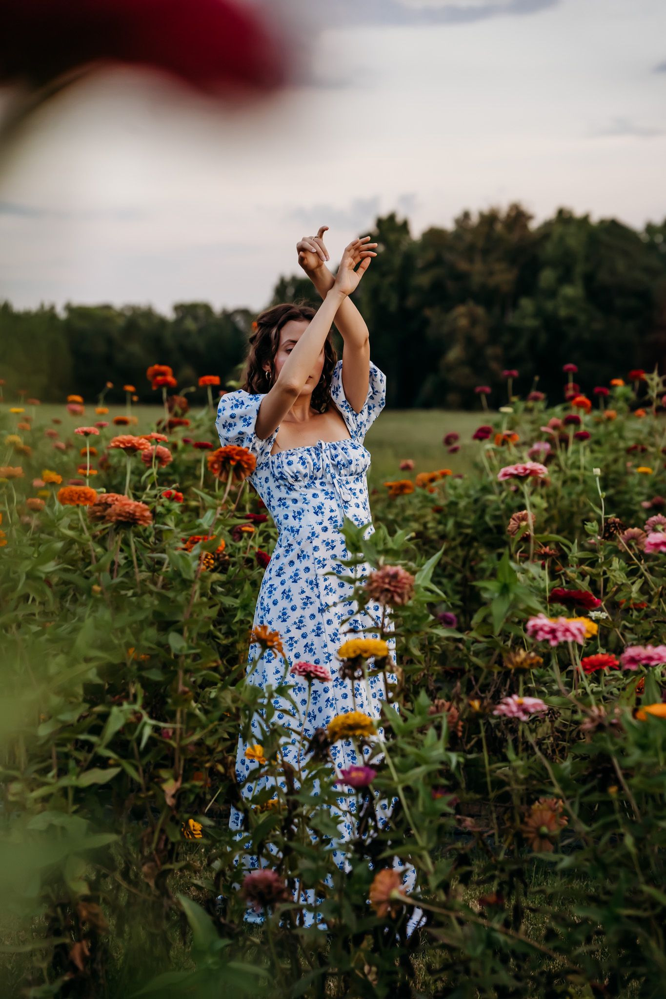 Woman in blue floral dress raising arms among colorful zinnia blooms in garden field.