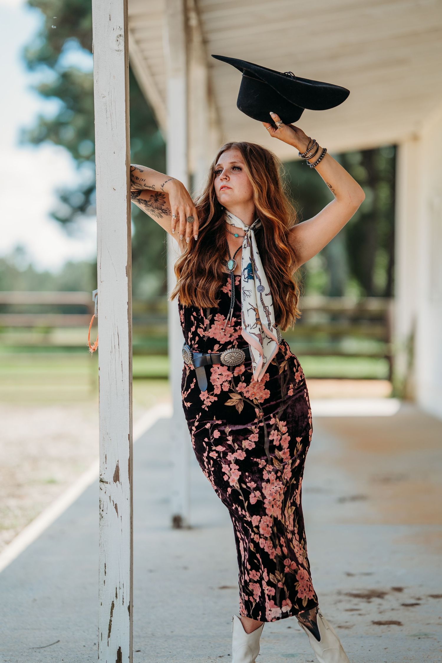 Woman in floral dress holding cowboy hat posing against white post at ranch.