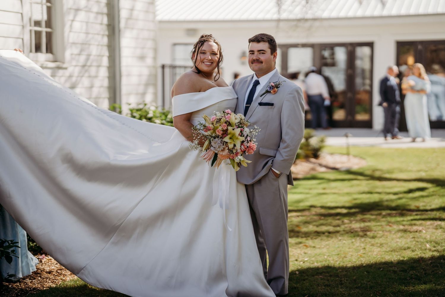 Smiling bride and groom pose outdoors with flowing white dress and colorful bridal bouquet.