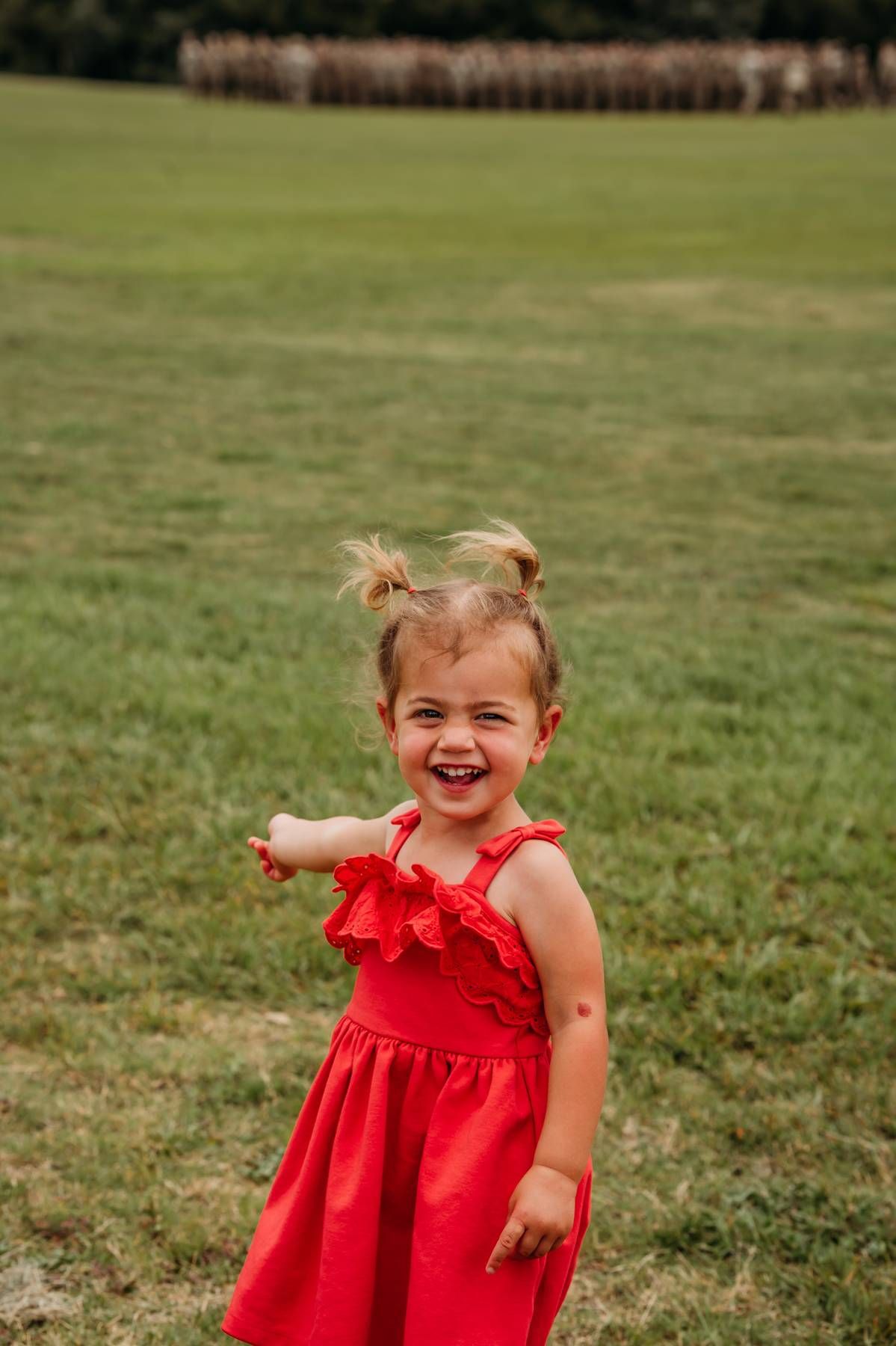 Joyful toddler girl in red dress with pigtails laughing outdoors on green grass field.