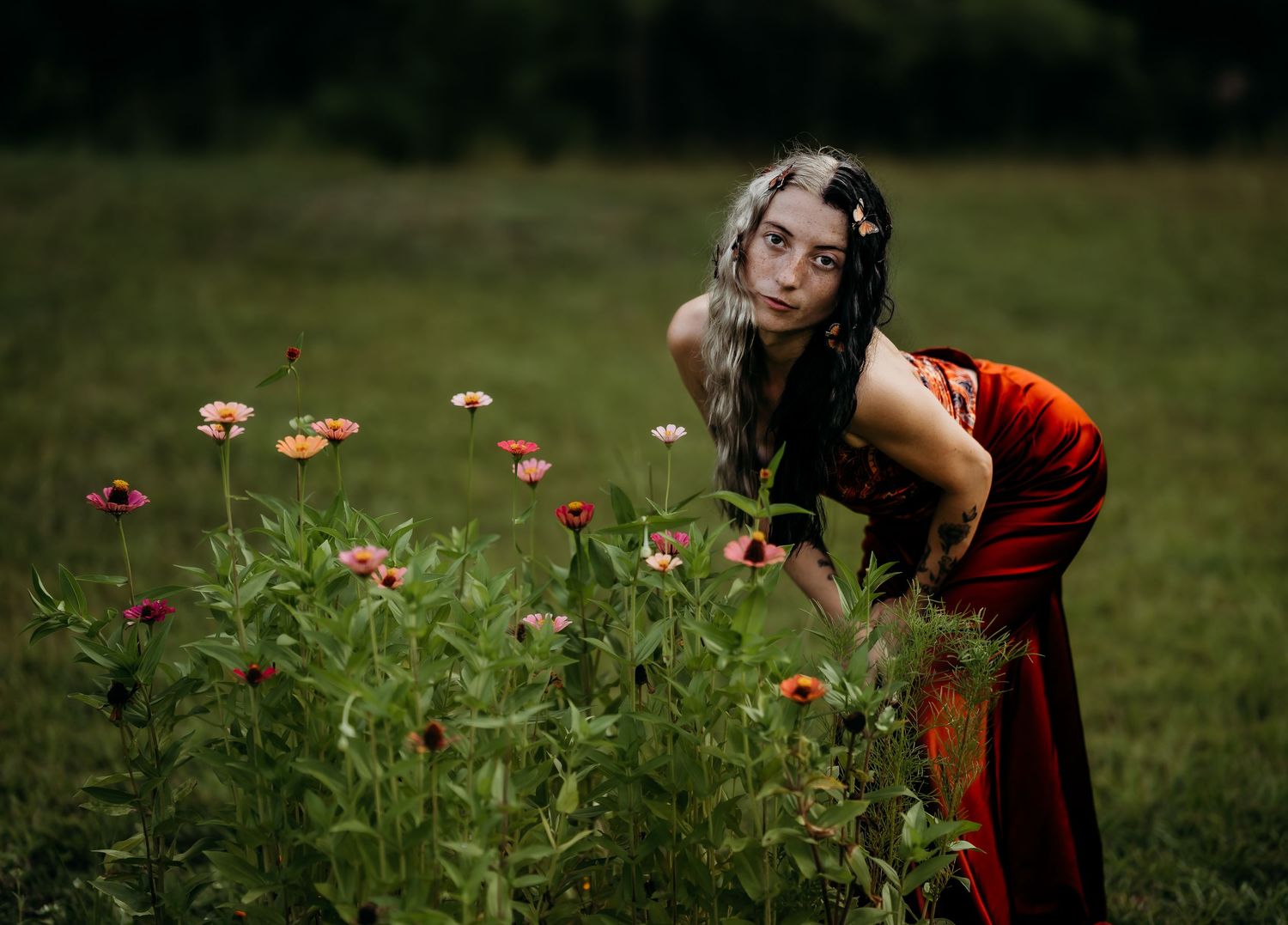 A young woman in a red dress leans over colorful zinnia flowers in a lush green field.
