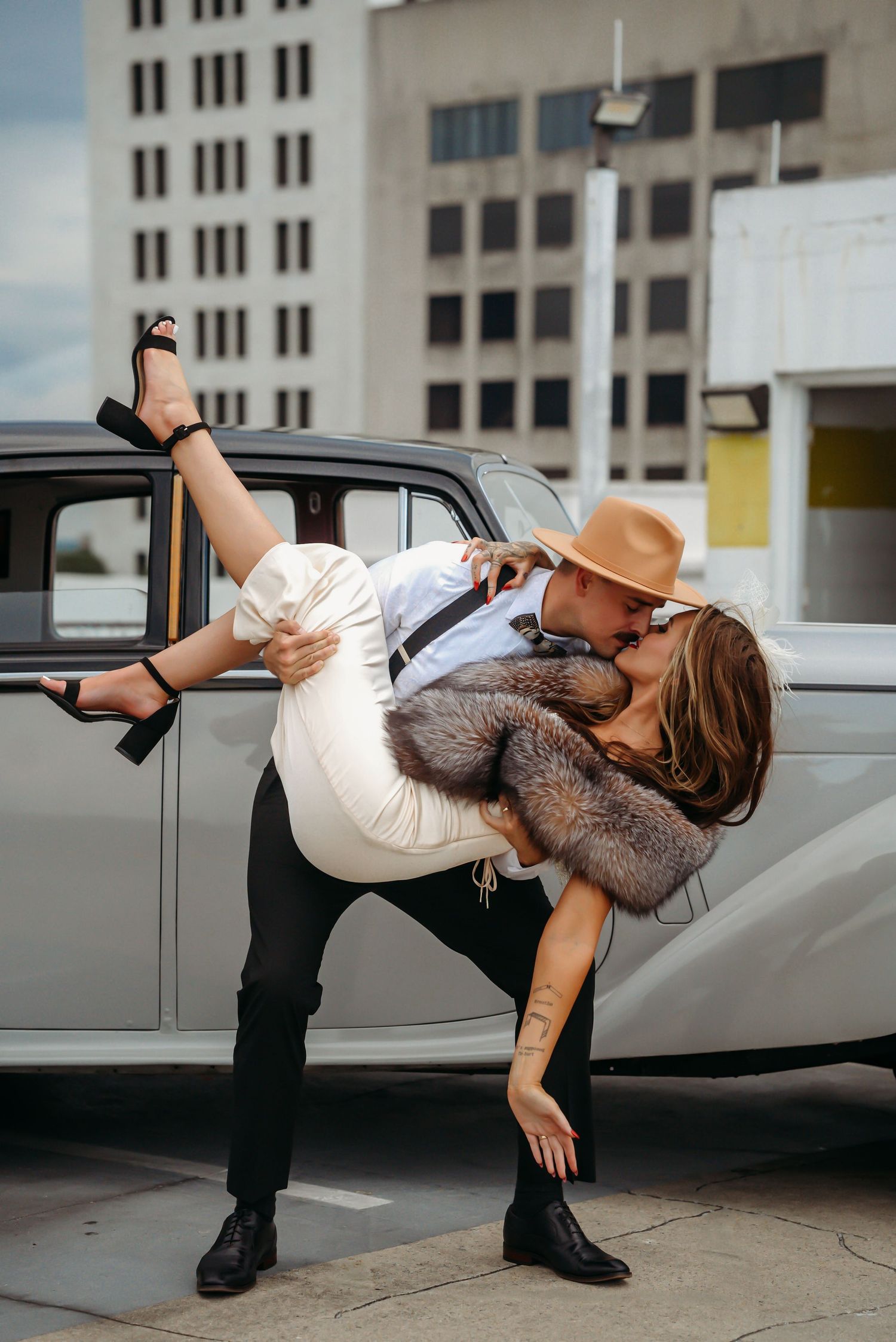 Couple sharing a romantic dip kiss beside a vintage white car, woman in fur coat and heels, man in hat and suspenders.