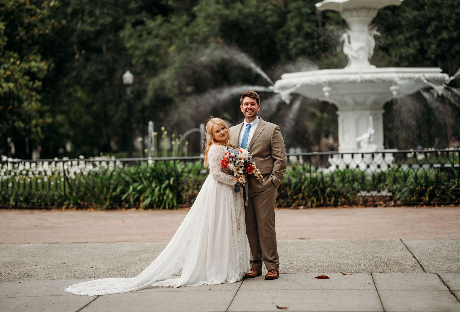 Wedding couple stands together near ornate white fountain, bride in lace gown holding vibrant bouquet.