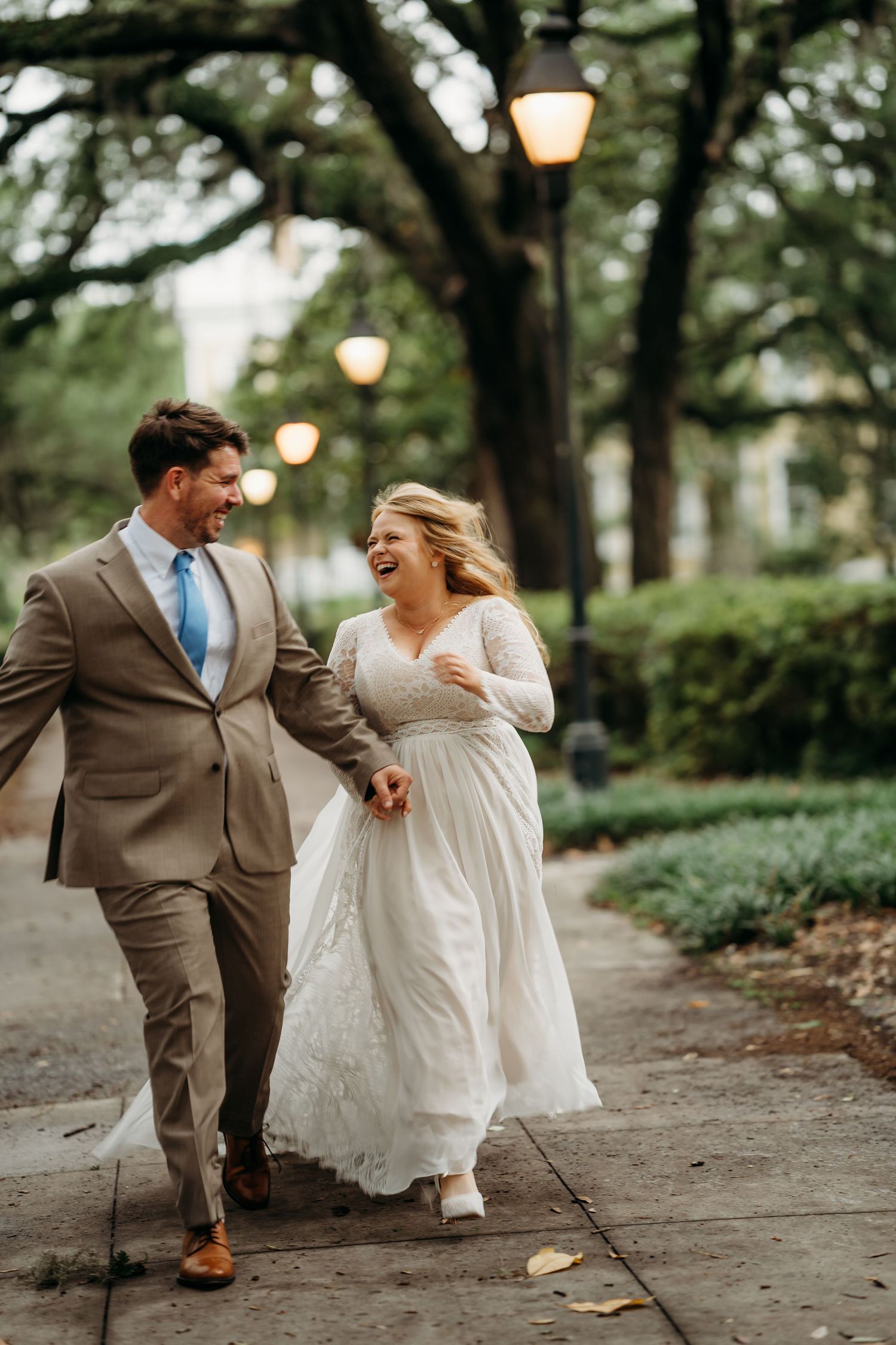 Laughing bride in white lace gown and groom in tan suit walk hand-in-hand along a tree-lined path with glowing lanterns.
