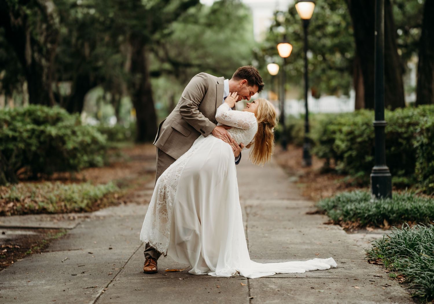 Newlyweds share a tender dip kiss along a scenic tree-lined path with glowing lamp posts.