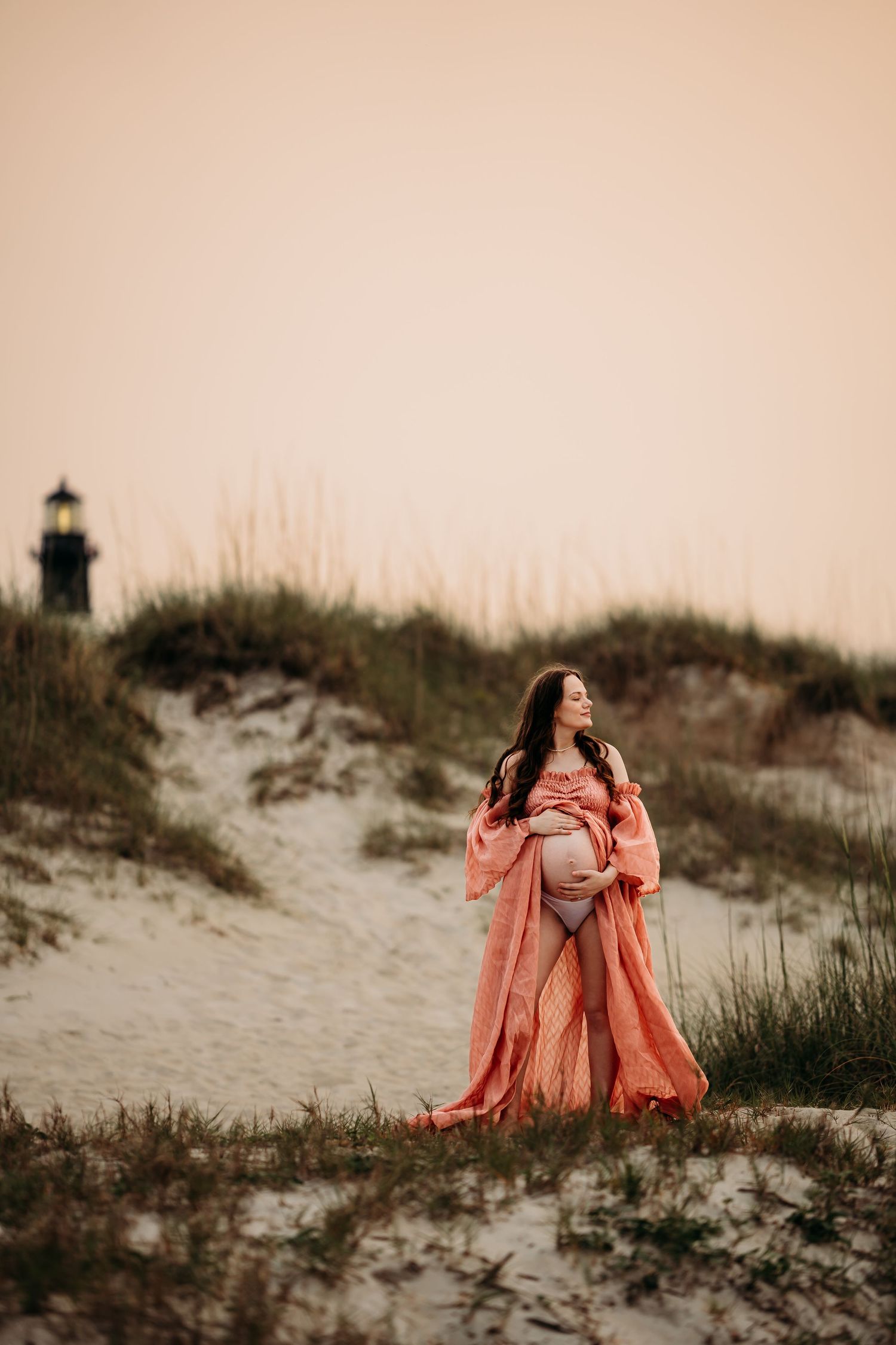 Pregnant woman in coral maternity gown standing on beach dunes with lighthouse in background at dusk.