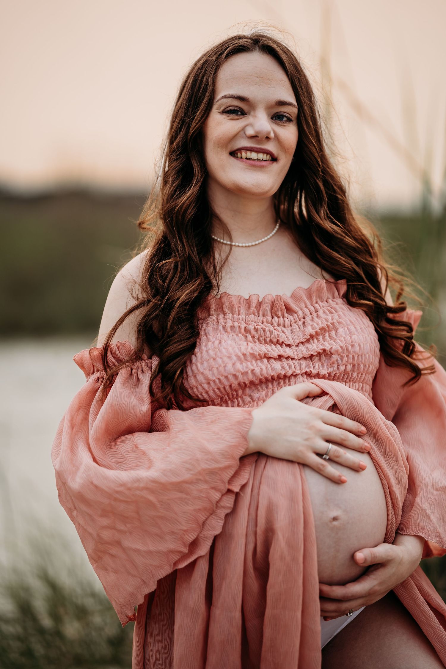 Smiling pregnant woman in coral off-shoulder maternity gown holding baby bump outdoors at sunset.