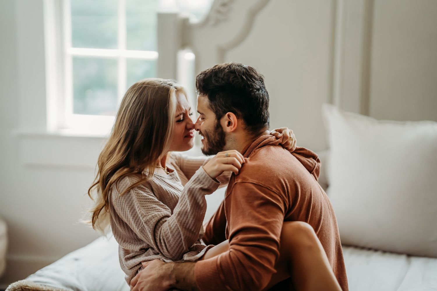 Happy couple sitting on a bed, faces close together, smiling and holding hands warmly.