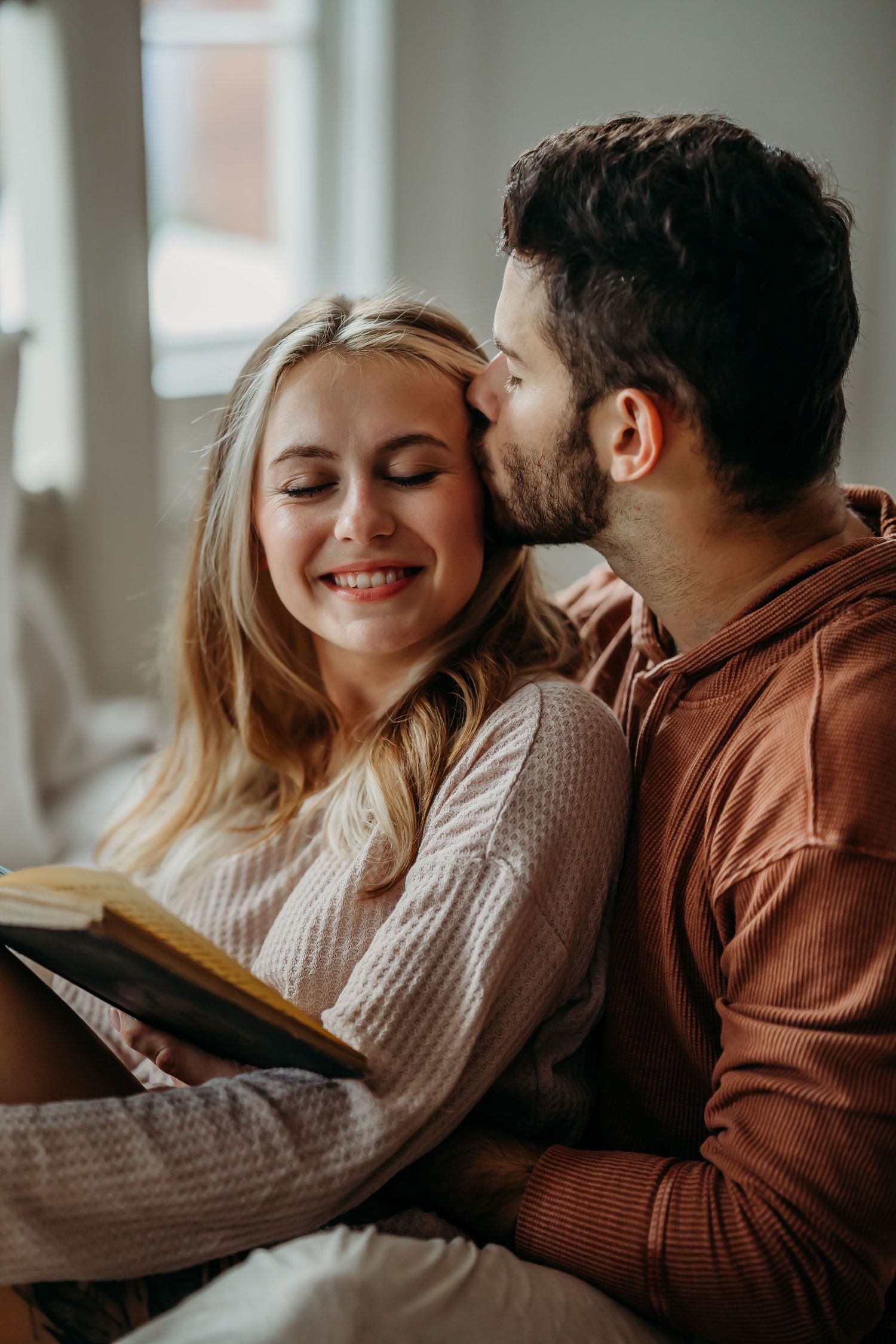 Man kissing smiling blonde woman on forehead while she reads book in bed.