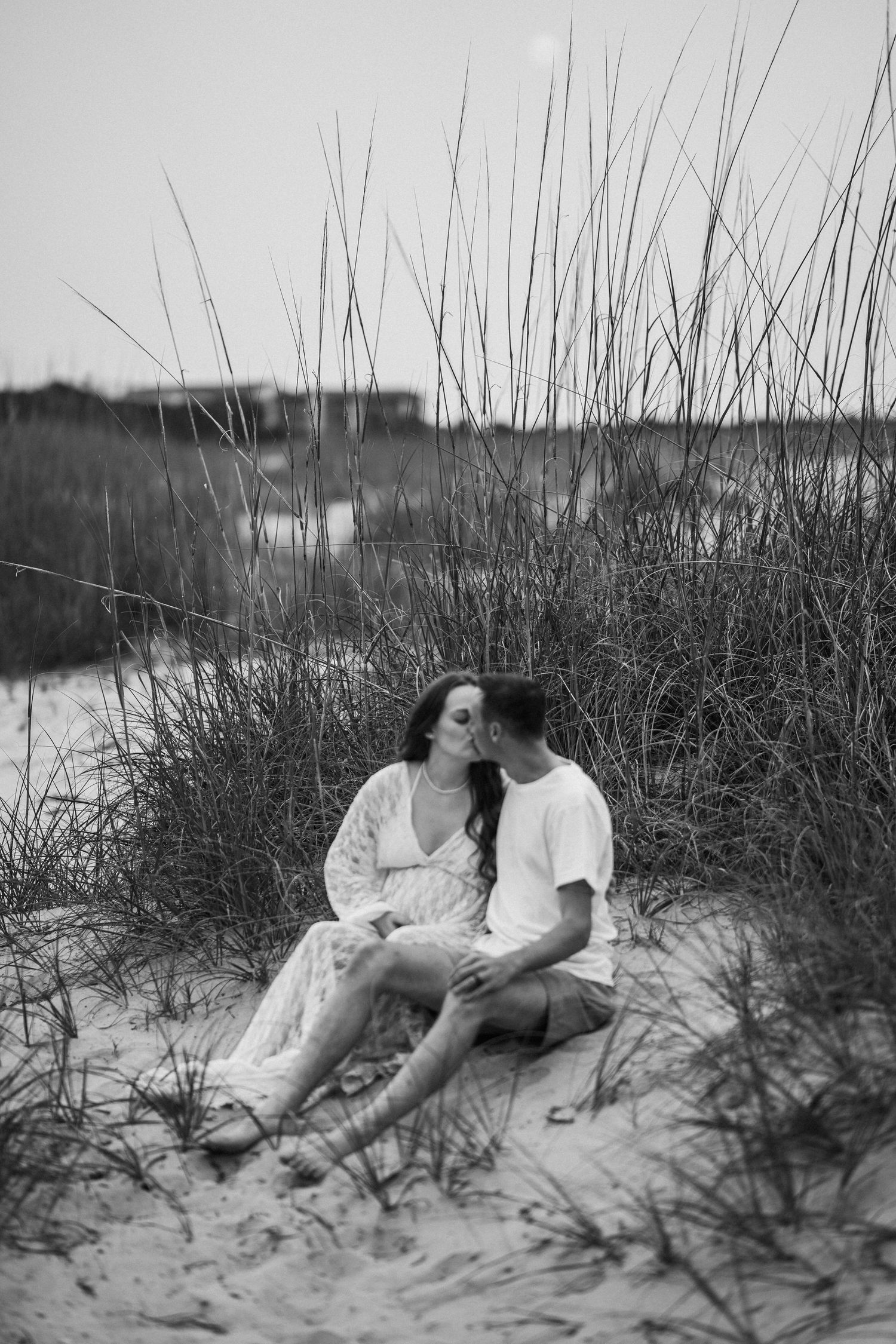 Couple sharing a kiss while sitting on sandy beach dunes surrounded by tall sea grass in black and white.