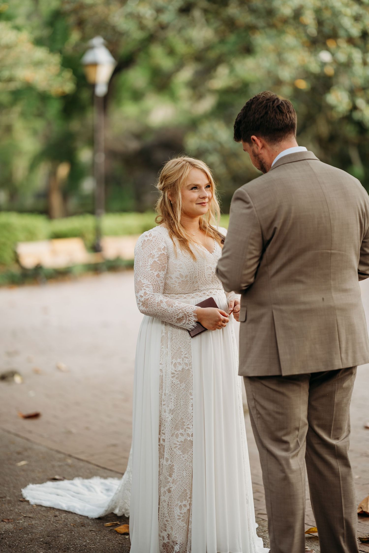Bride in lace wedding gown gazes at groom while exchanging vows in a lush park setting.