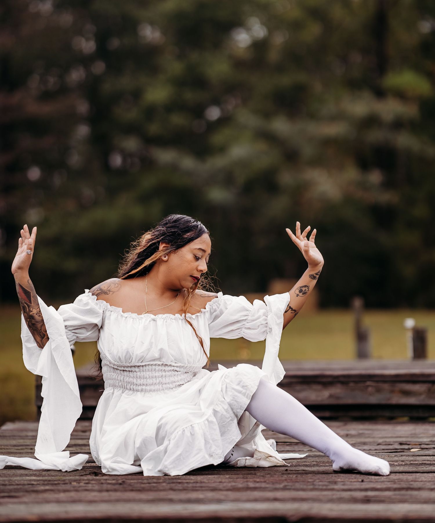 Tattooed woman in white smocked dress poses expressively seated on a rustic wooden dock.