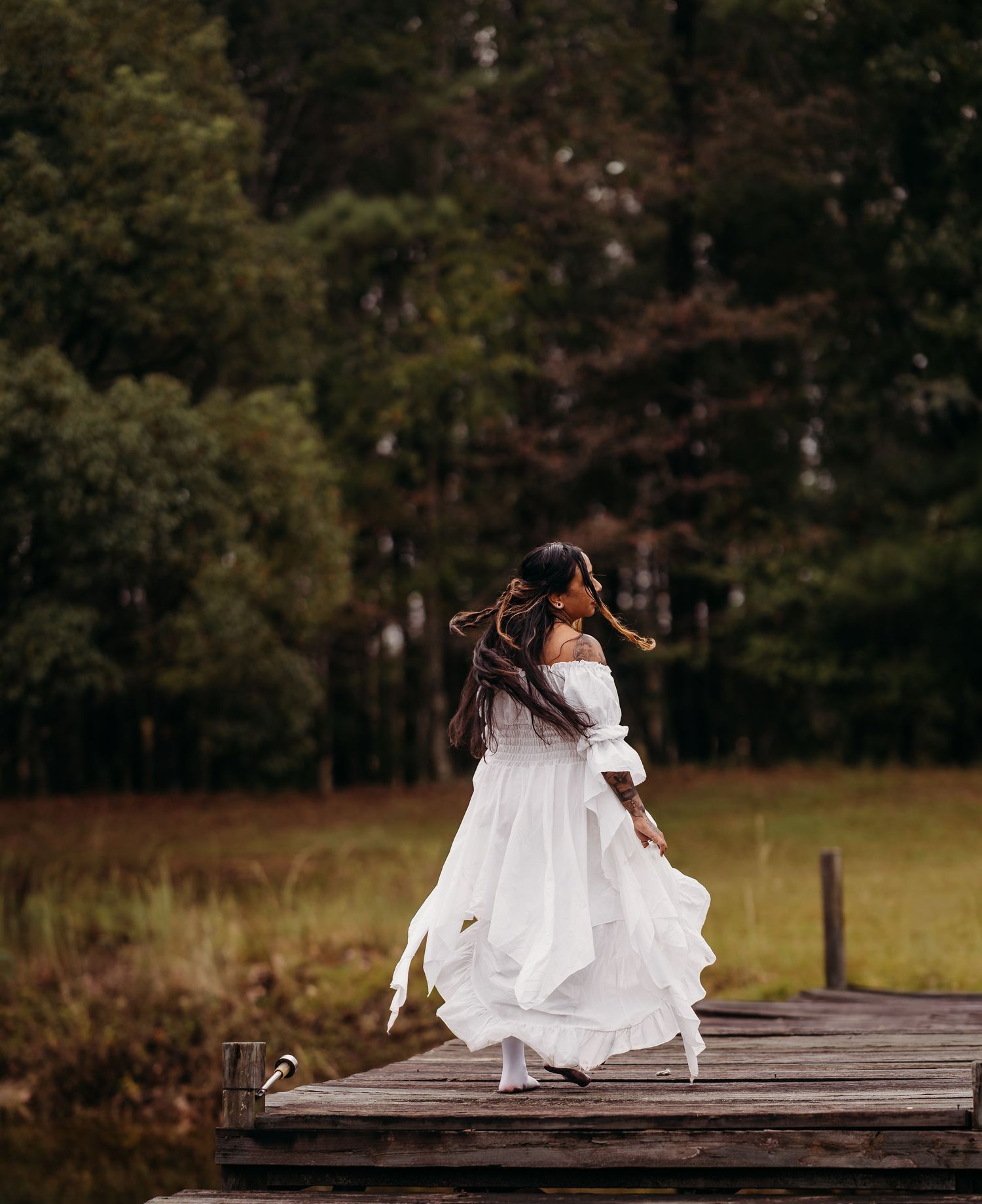 Woman in white off-shoulder dress spinning on wooden dock with lush green forest background.