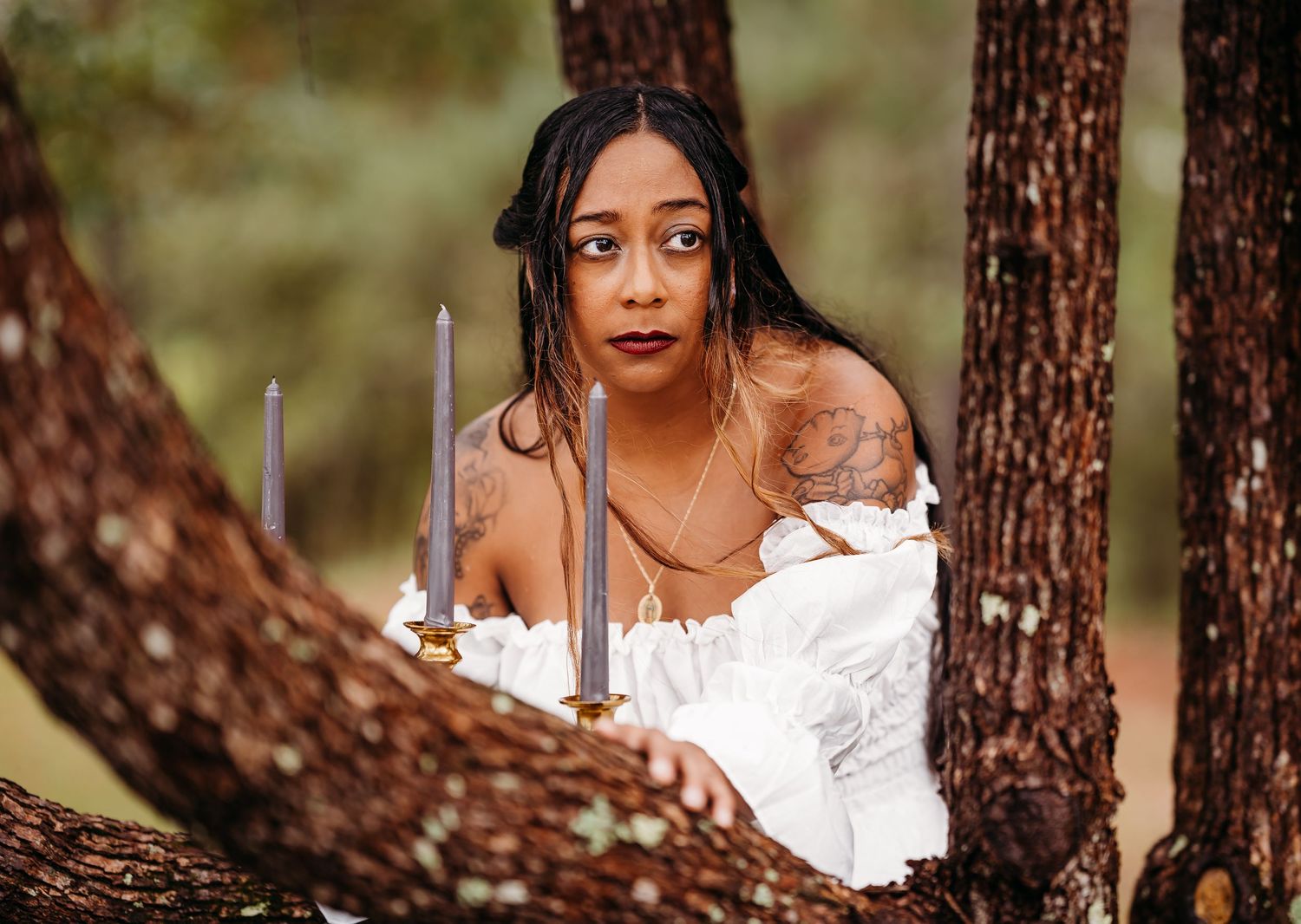 Woman in white off-shoulder dress holding candles between forest trees.