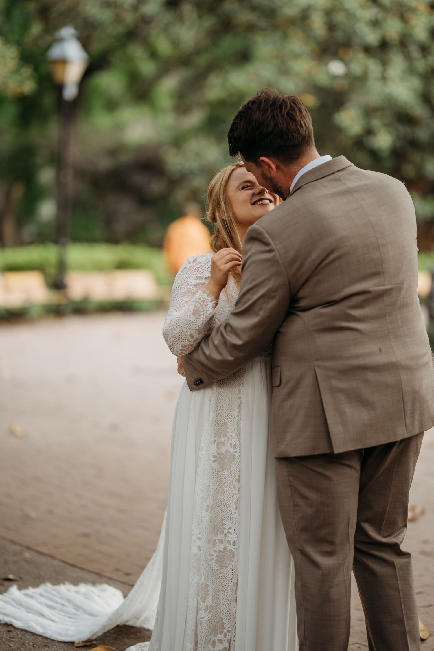 Bride in lace gown laughs joyfully as groom in tan suit embraces her in a lush green park setting.