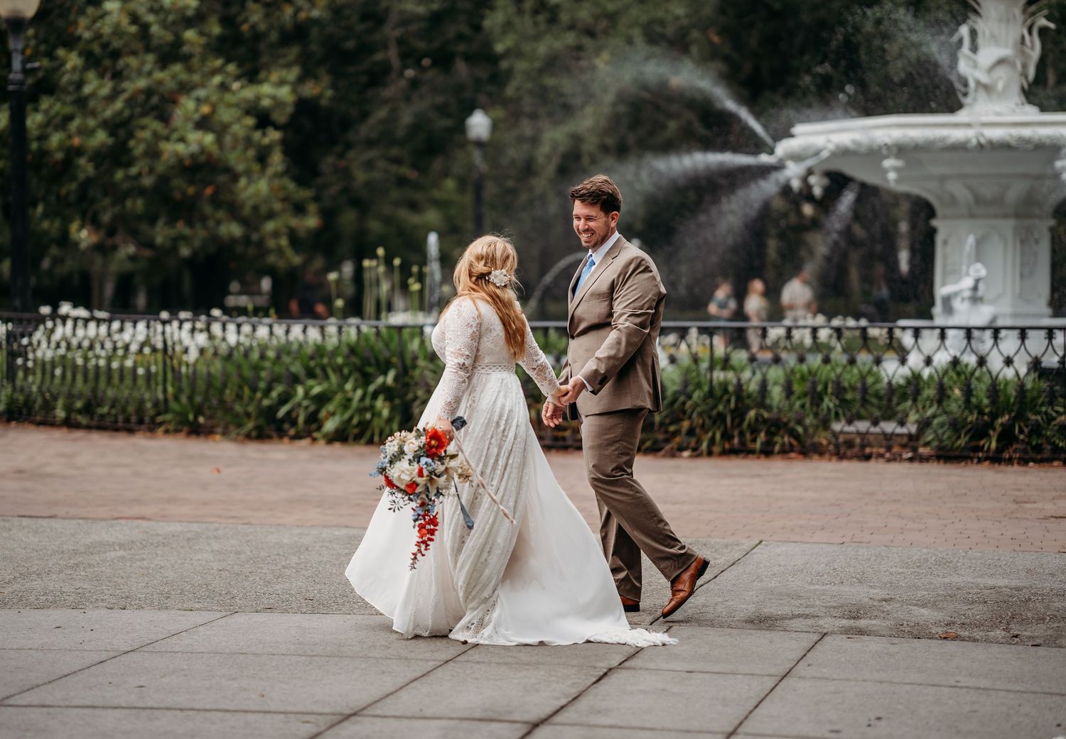 Bride and groom walk hand-in-hand past a white fountain in a park on their wedding day.