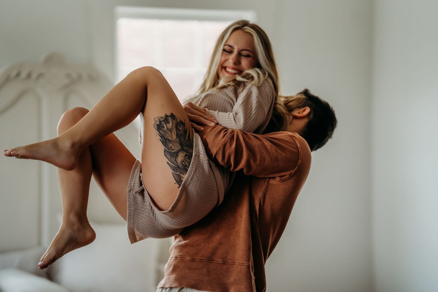 Man lifting tattooed laughing woman in his arms in bright bedroom.
