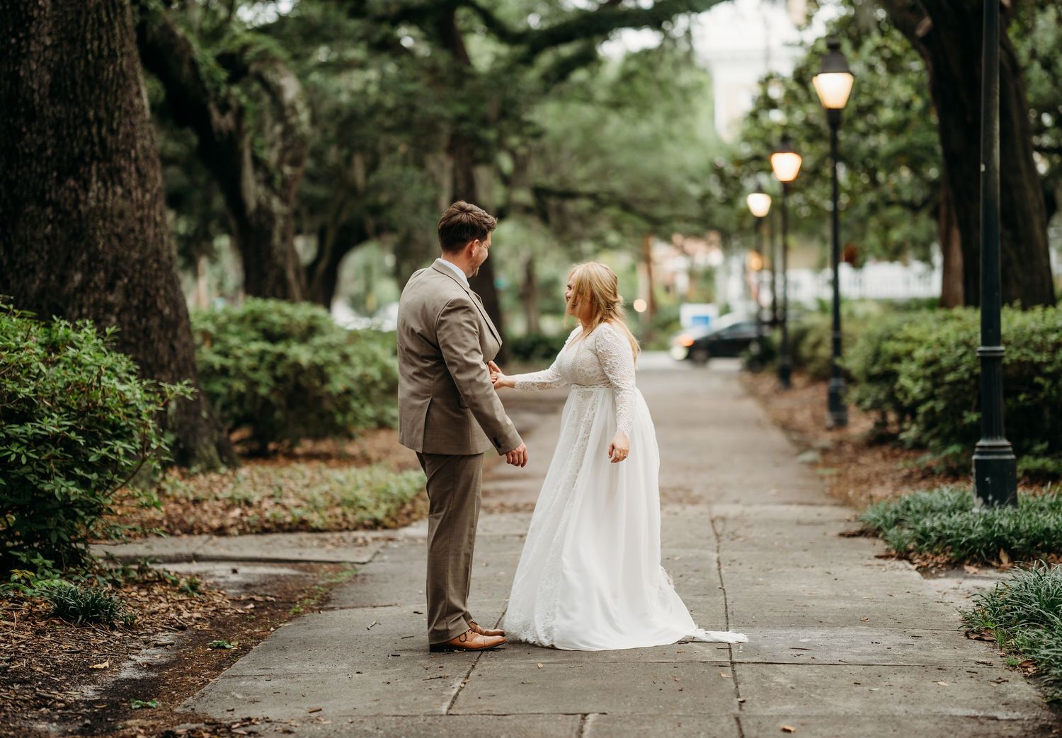 Bride in elegant white gown and groom in tan suit hold hands on a romantic tree-lined sidewalk with soft lamp light.