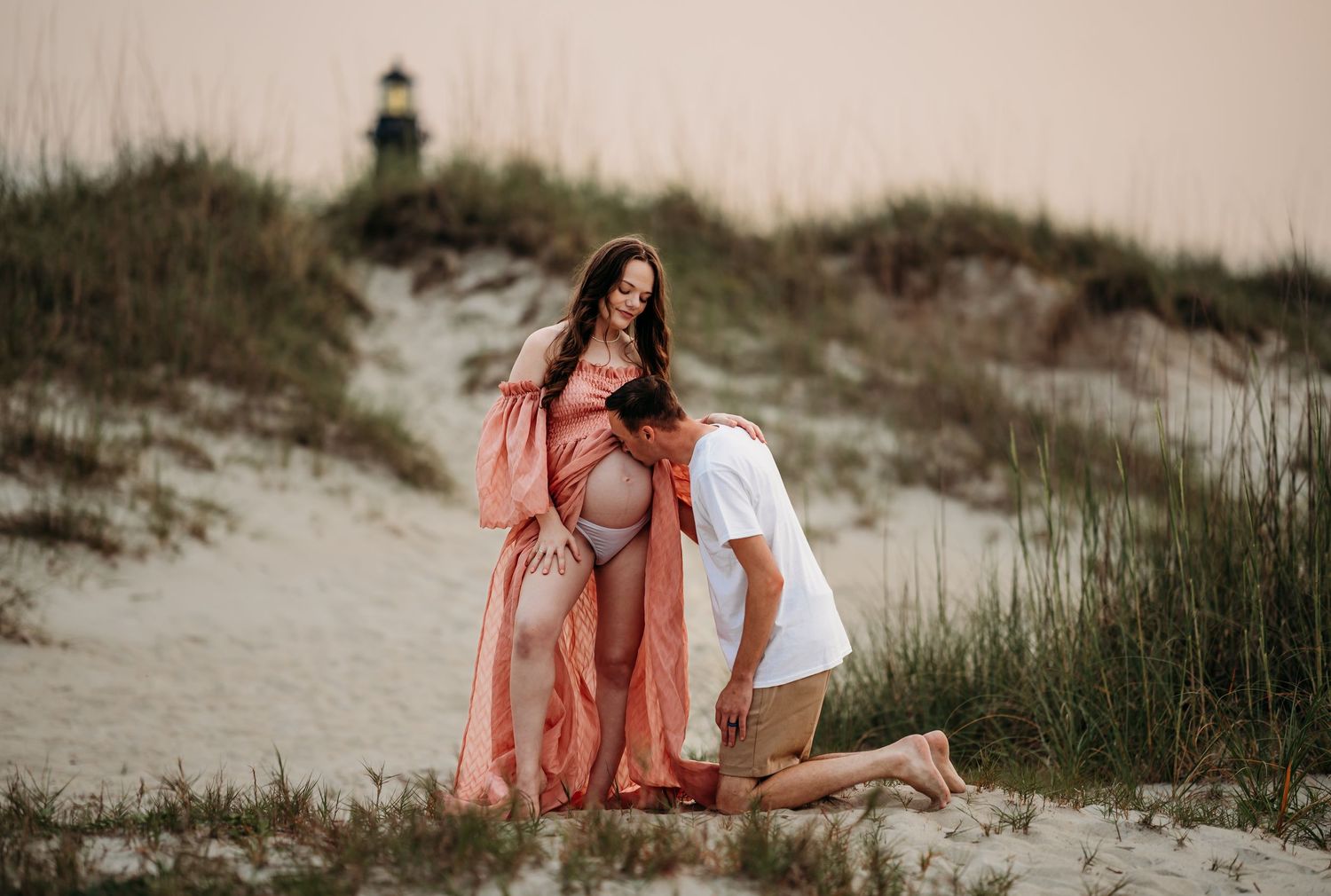 Man kneeling before pregnant partner on beach dunes with lighthouse backdrop, woman in coral gown.