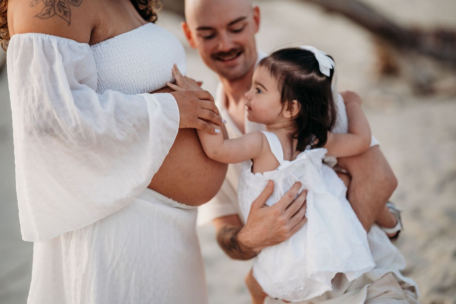 A pregnant woman in a white dress with her partner and toddler daughter on a beach during a maternity photoshoot.