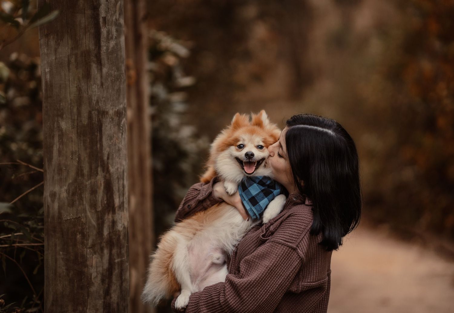 A woman with dark hair kisses her fluffy Pomeranian dog outdoors on an autumn forest path.