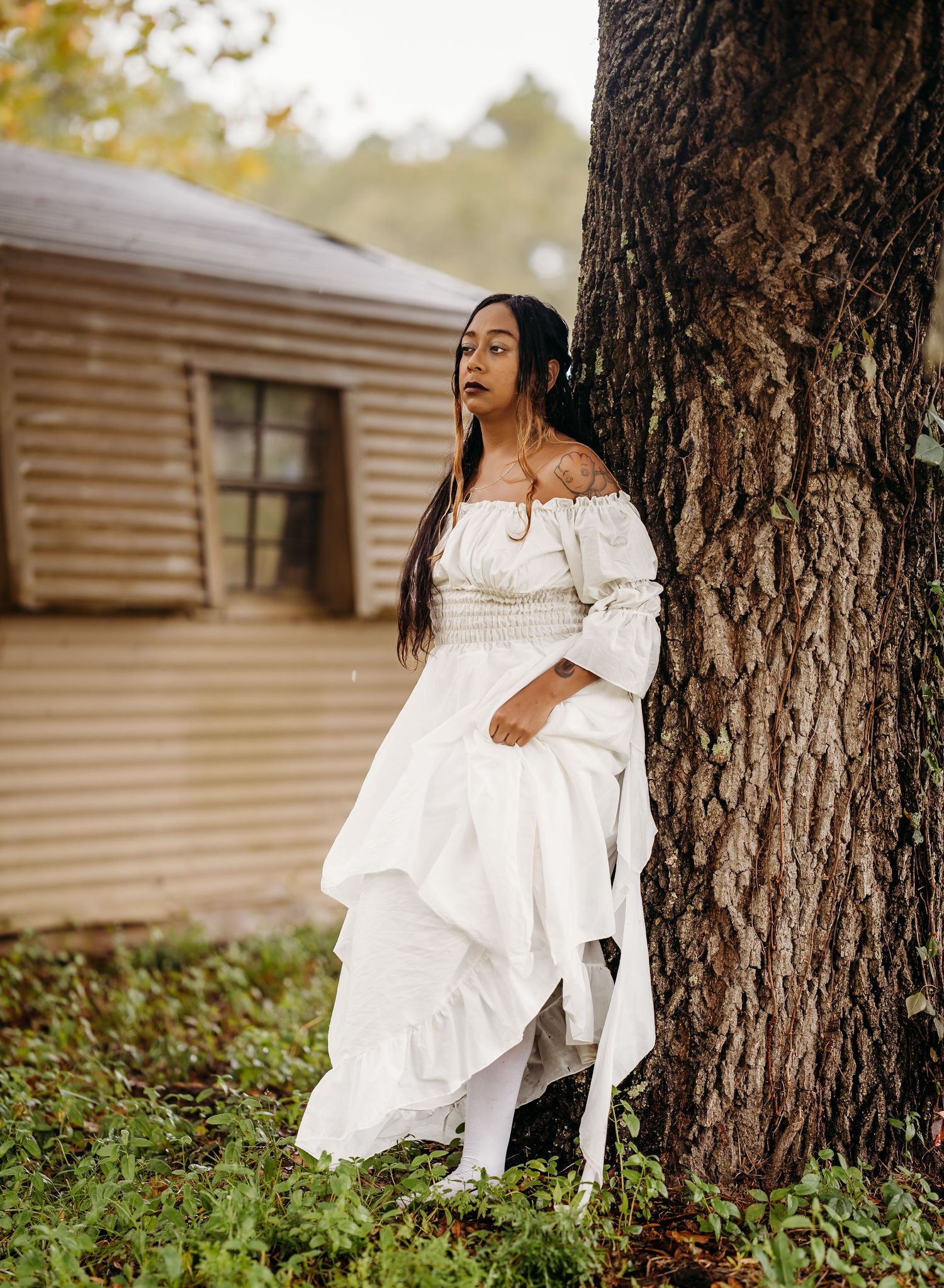 Woman in white ruffled off-shoulder gown leaning against a tree beside a wooden cabin.
