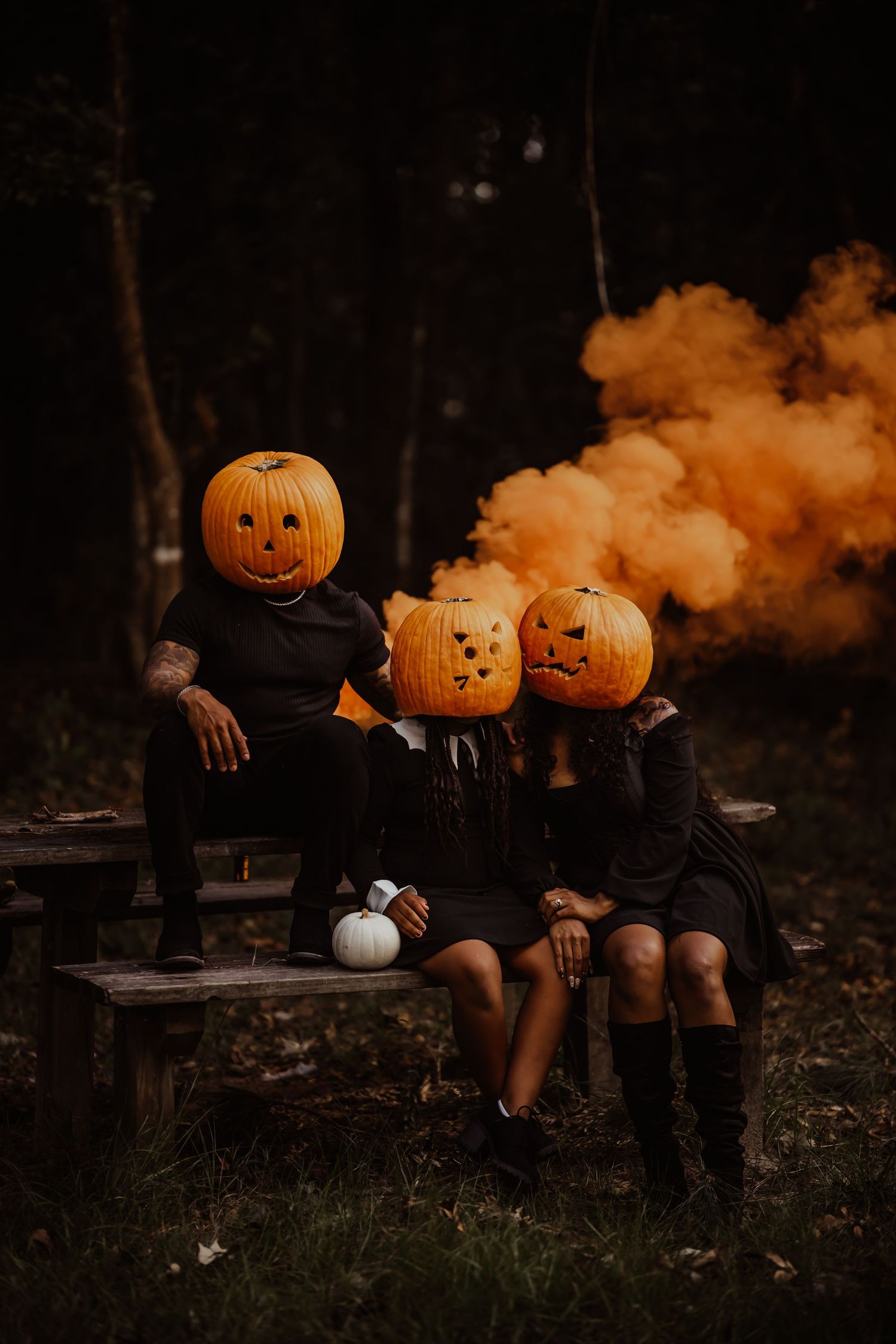 Two people wearing carved jack-o-lantern pumpkin heads sit on bench with orange smoke in dark forest.