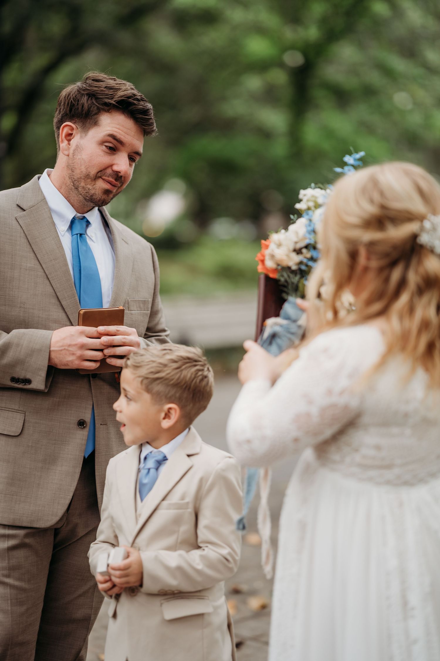 Groom in tan suit reading vows as ring bearer and bride look on outdoors.