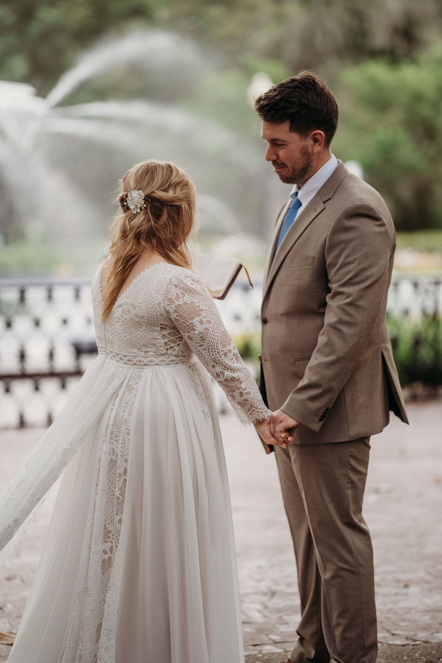 Bride in lace gown and groom in tan suit hold hands tenderly before a misty garden fountain.
