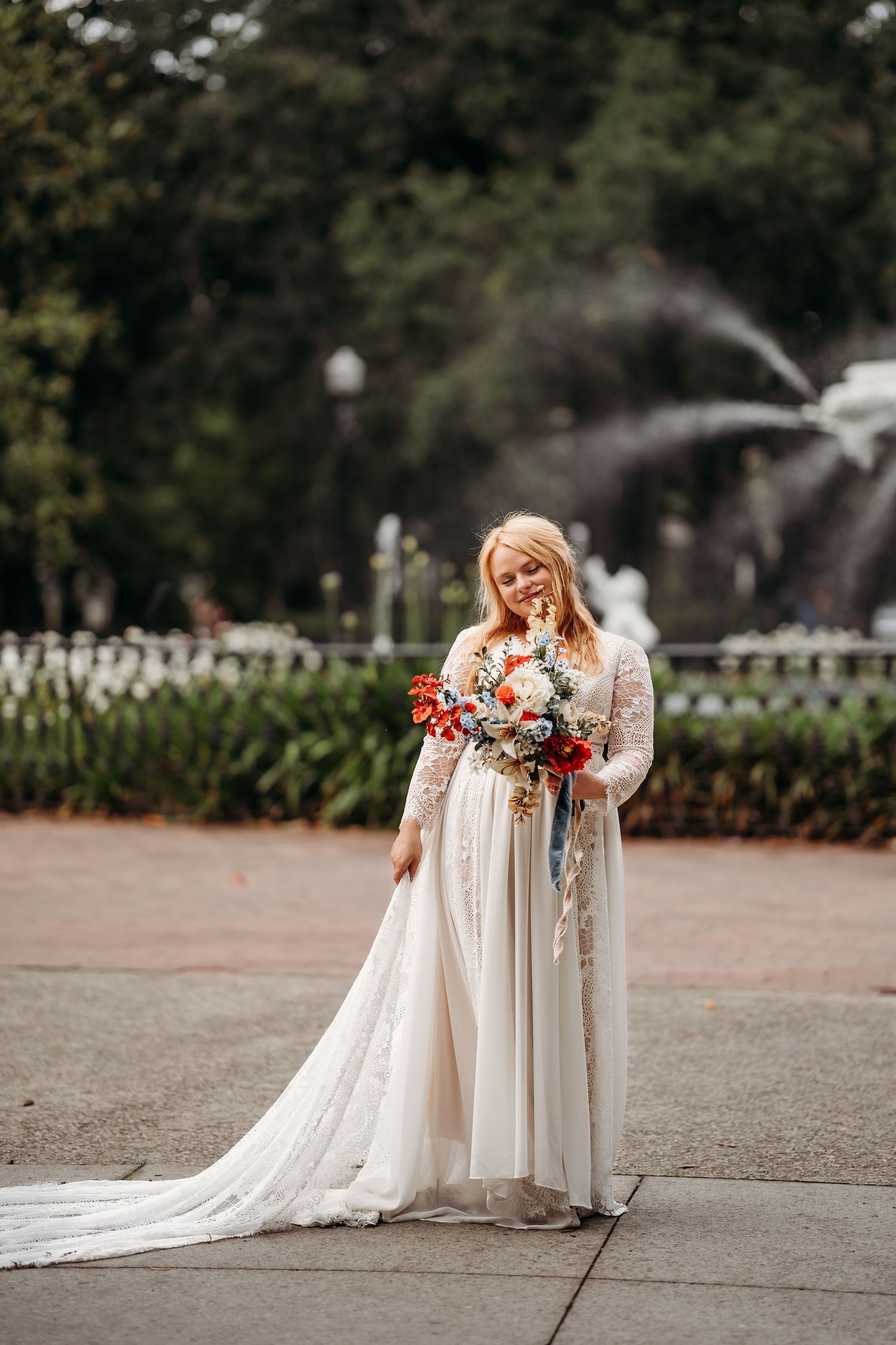 Bride in lace wedding gown smiles down at colorful bouquet with fountain in background.