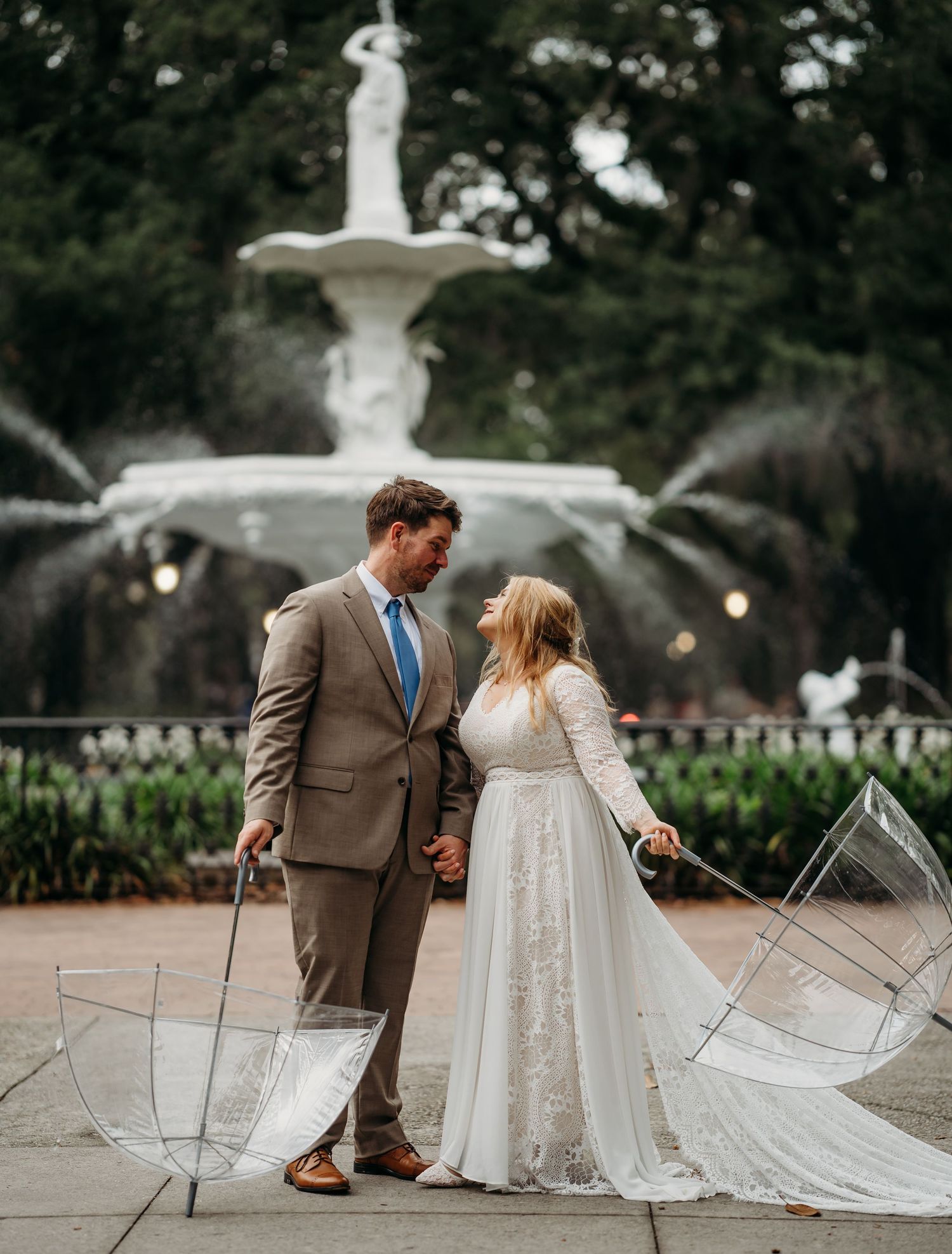 Bride and groom hold hands gazing at each other with clear umbrellas before ornate white fountain.