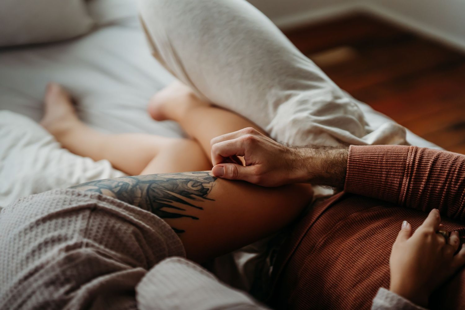 Intimate close-up of tattooed man gently touching partner's leg on cozy bed.
