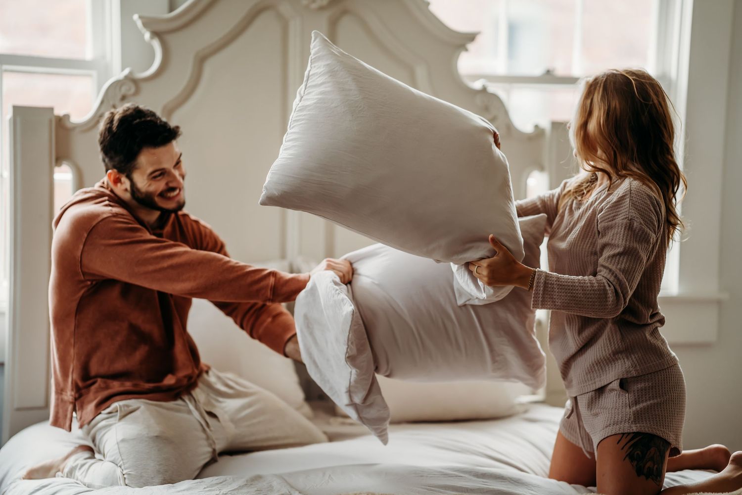A happy couple having a playful pillow fight on a white bed in a bright, elegant bedroom.