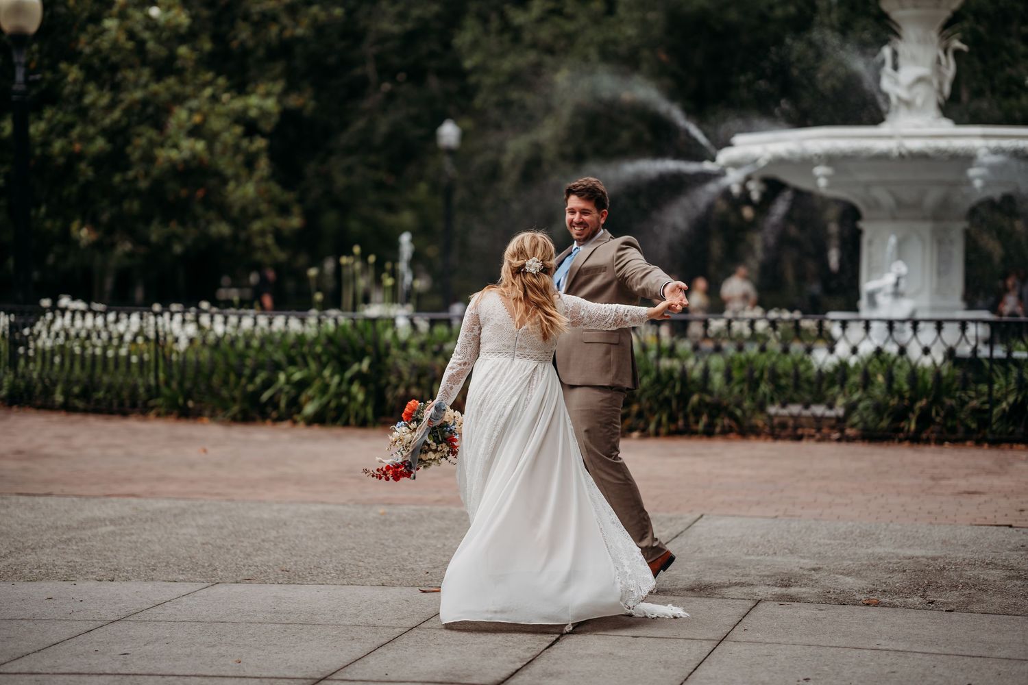 Bride and groom dance together near a white fountain, her lace gown and bouquet swirling as they spin.