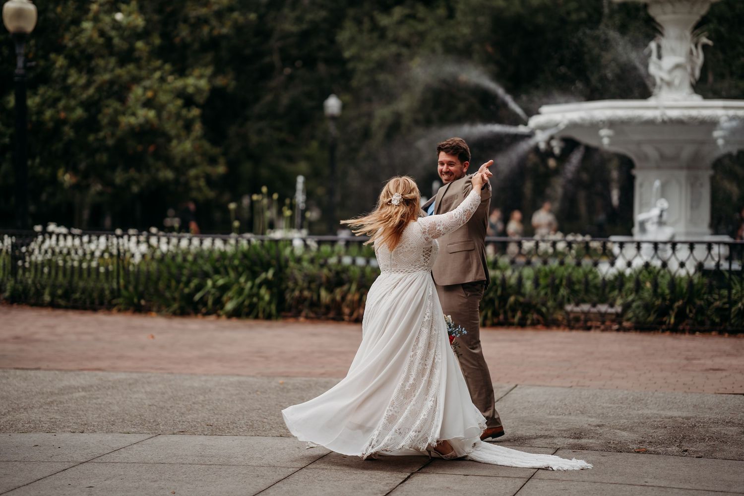Bride and groom dance joyfully in front of a white fountain, her lace gown flowing gracefully in the breeze.