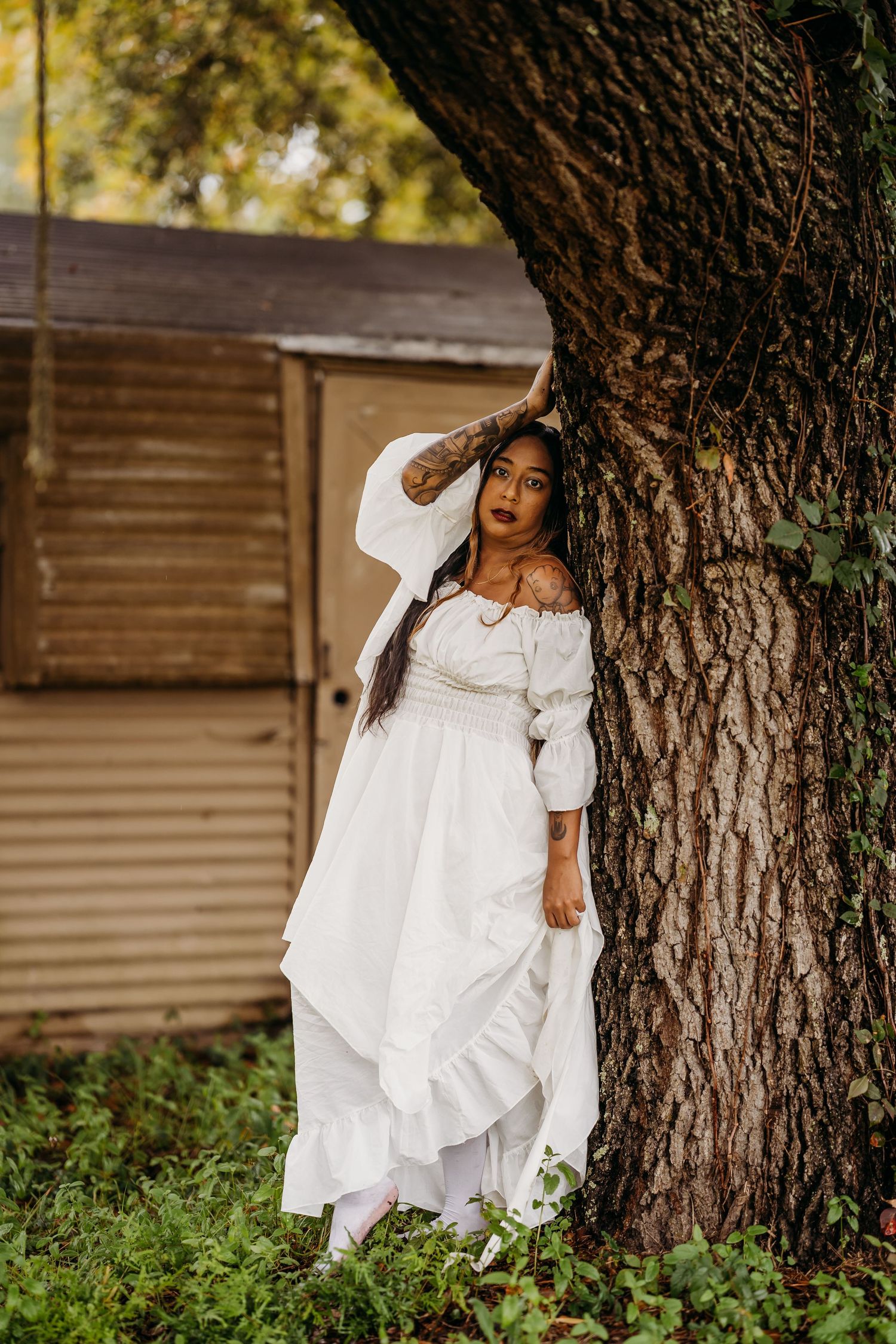 Woman in white off-shoulder dress leaning against large tree trunk near rustic wooden shed.