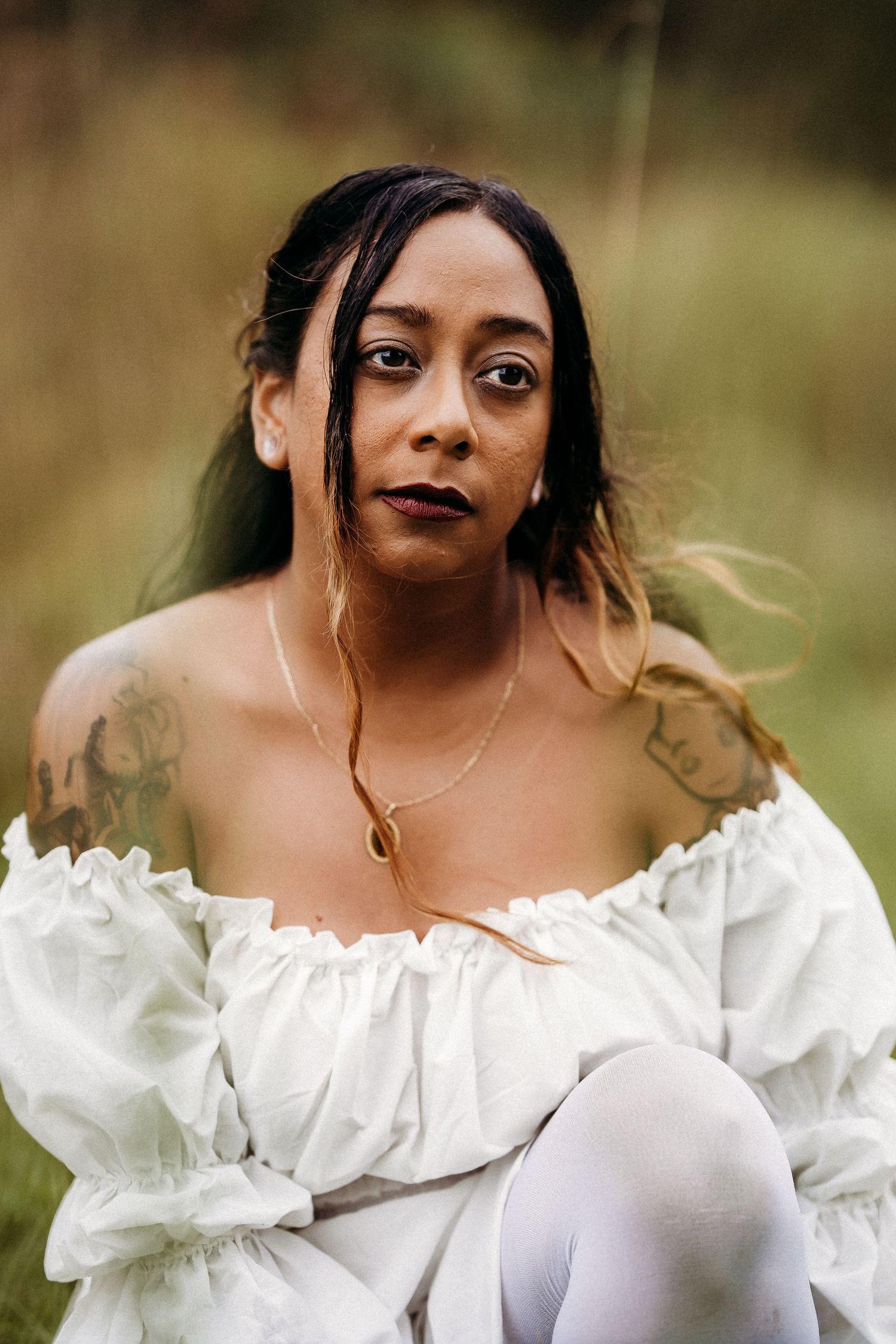 Close-up portrait of tattooed woman in white off-shoulder dress sitting outdoors in nature.