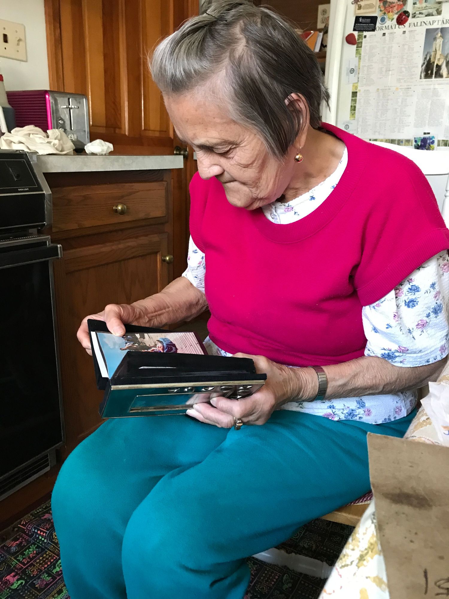 Elderly woman in red top sitting in kitchen looking at photo album fondly.