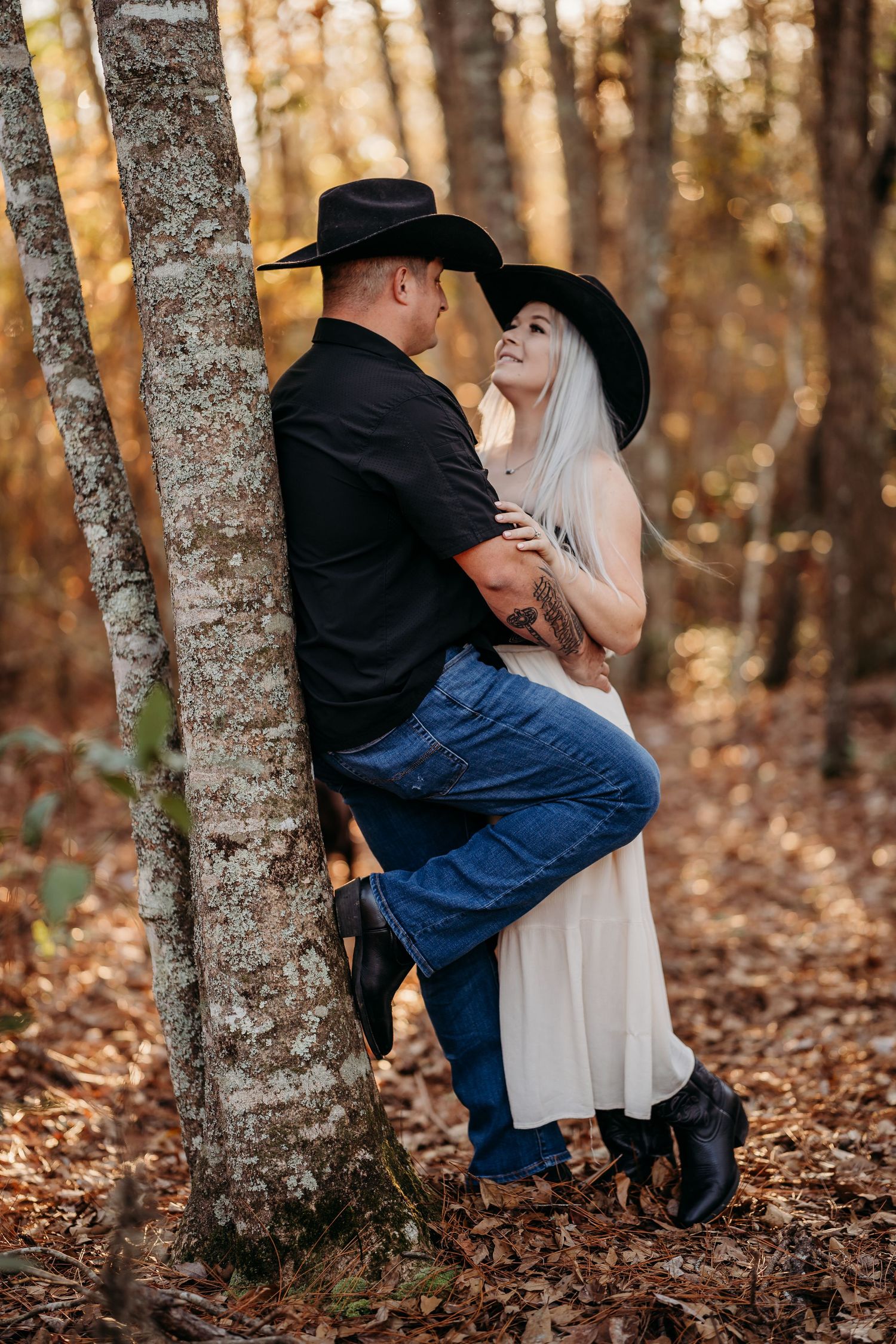 Couple in black cowboy hats embracing by a birch tree in an autumn forest, wearing jeans and a white dress.